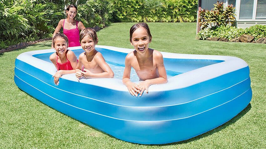 Blauer viereckiger aufgeblasener Pool in dem 3 Kinder spielen. Daneben steht eine Frau und passt auf.